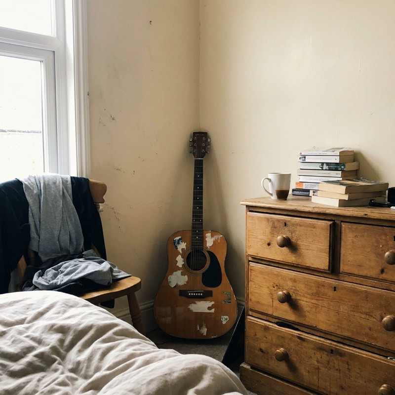 A worn acoustic guitar propped in a bedroom corner.